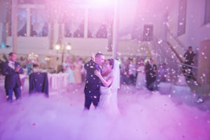 A newly wed couple dance together amidst colored smoke, confetti, and soft pink lighting.