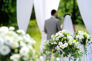 A beautiful flower arrangement is shown in the foreground while a couple getting married are shown in the background.